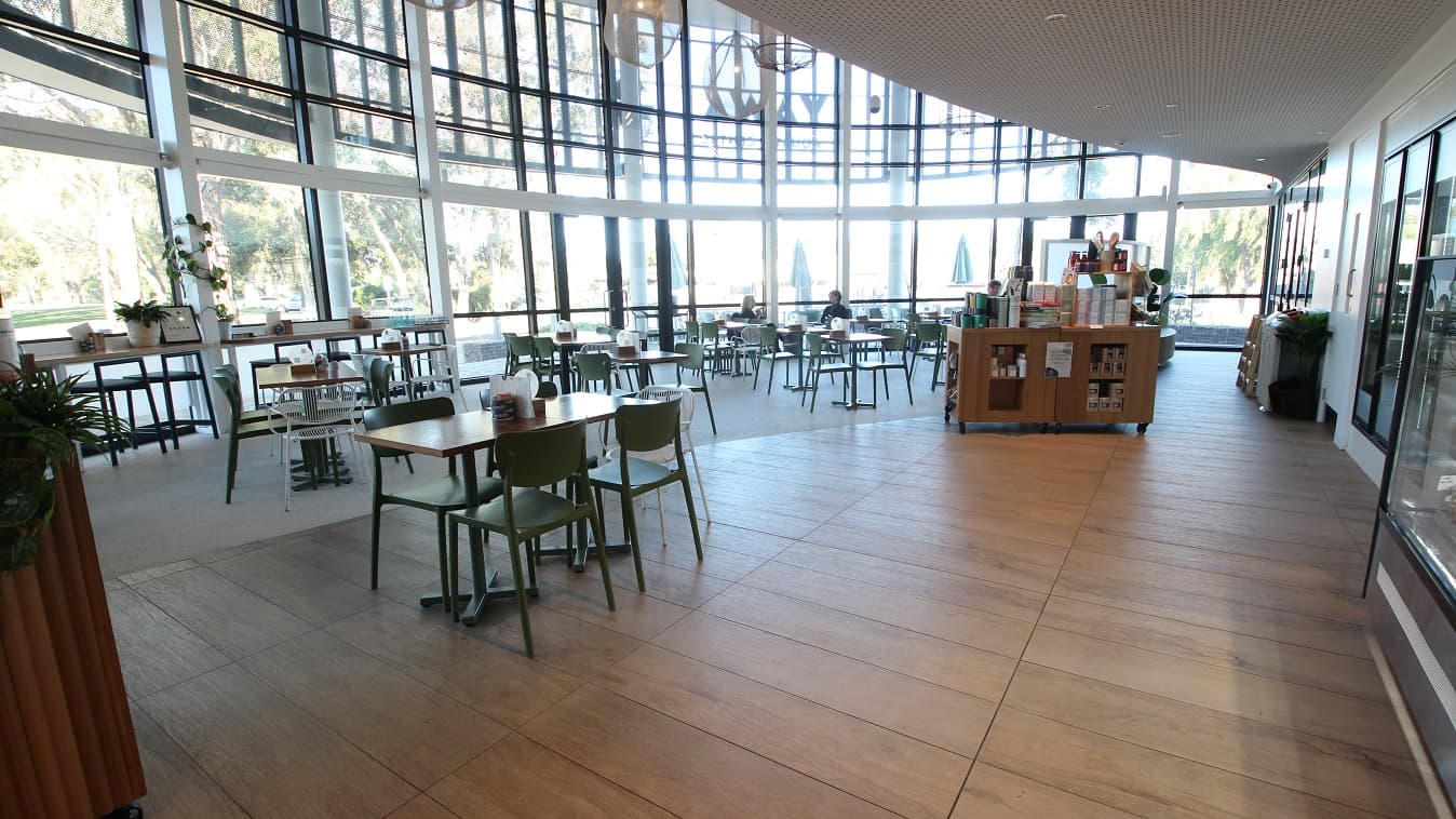 The café's seating area is illuminated by natural light coming in through the glass walls, and jelly-like artificial lights hanging from the ceiling provide additional lighting support. There is a narrow table with black high chairs located near the glass walls. A small cabinet that has tumblers and boxes is also available. Half of the seating area has an ash hardwood floor, while the other half is white.