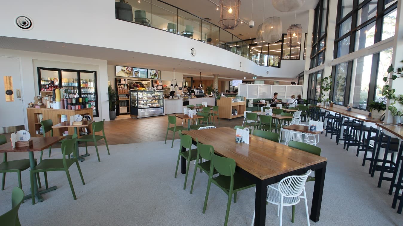 View of the entire area of Elements Eatery at Yawa Aquatic Centre. The café has flexible seating options, including easy-to-move white and green chairs and a space for wheelchairs and prams. There is a narrow table with black high chairs located near the glass walls. There is a green sofa chair before the panel board for the working space. A small cabinet that has tumblers and boxes can be found near the 3-door fridge.