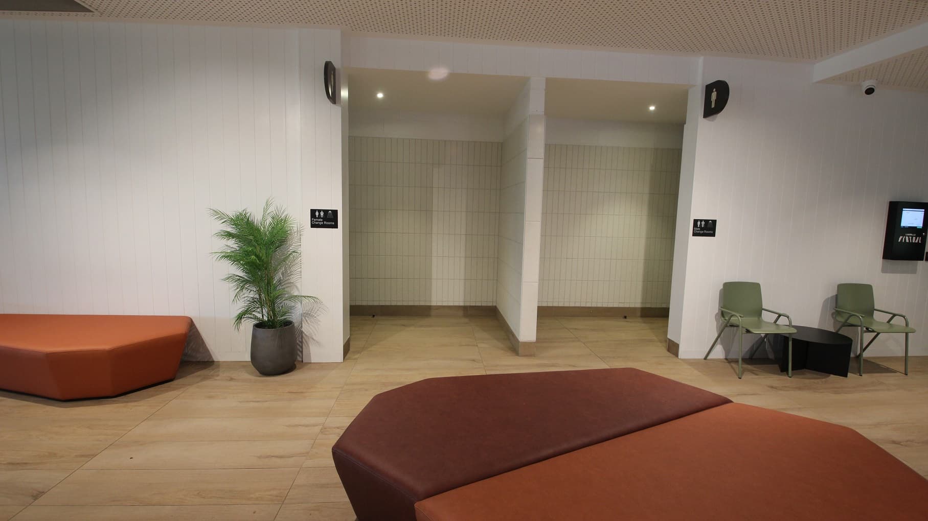The first floor at Yawa Aquatic Centre can be accessed by either stairs or via a lift. To the left of the lift are rubbish bins, a sanitizing dispenser, and a television mounted on the wall. To the right is the wooden stairwell with a metal handrail. From this position, we can see a glimpse of the Health Club through the glass walls.