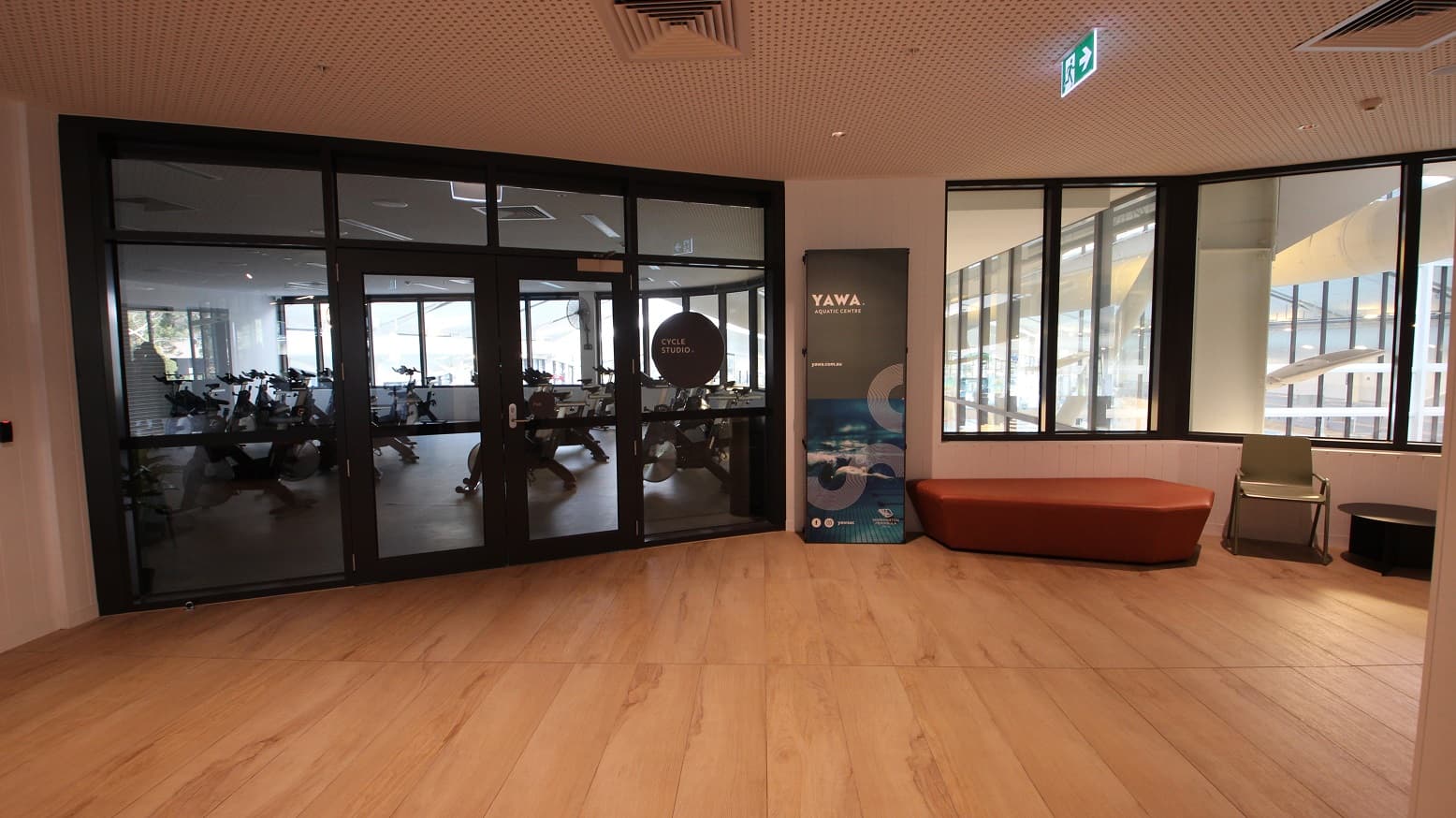 Cycle studio at Yawa Aquatic Centre. On the right is an orange flat sofa chair and a green armchair. The studio can be distinguished by the black circular sign board containing “Cycle studio” on the right glass door and the bicycles visible through the glass. Similar to the ground floor, this has an ash hardwood floor with a ceiling having small aesthetic holes as the design.