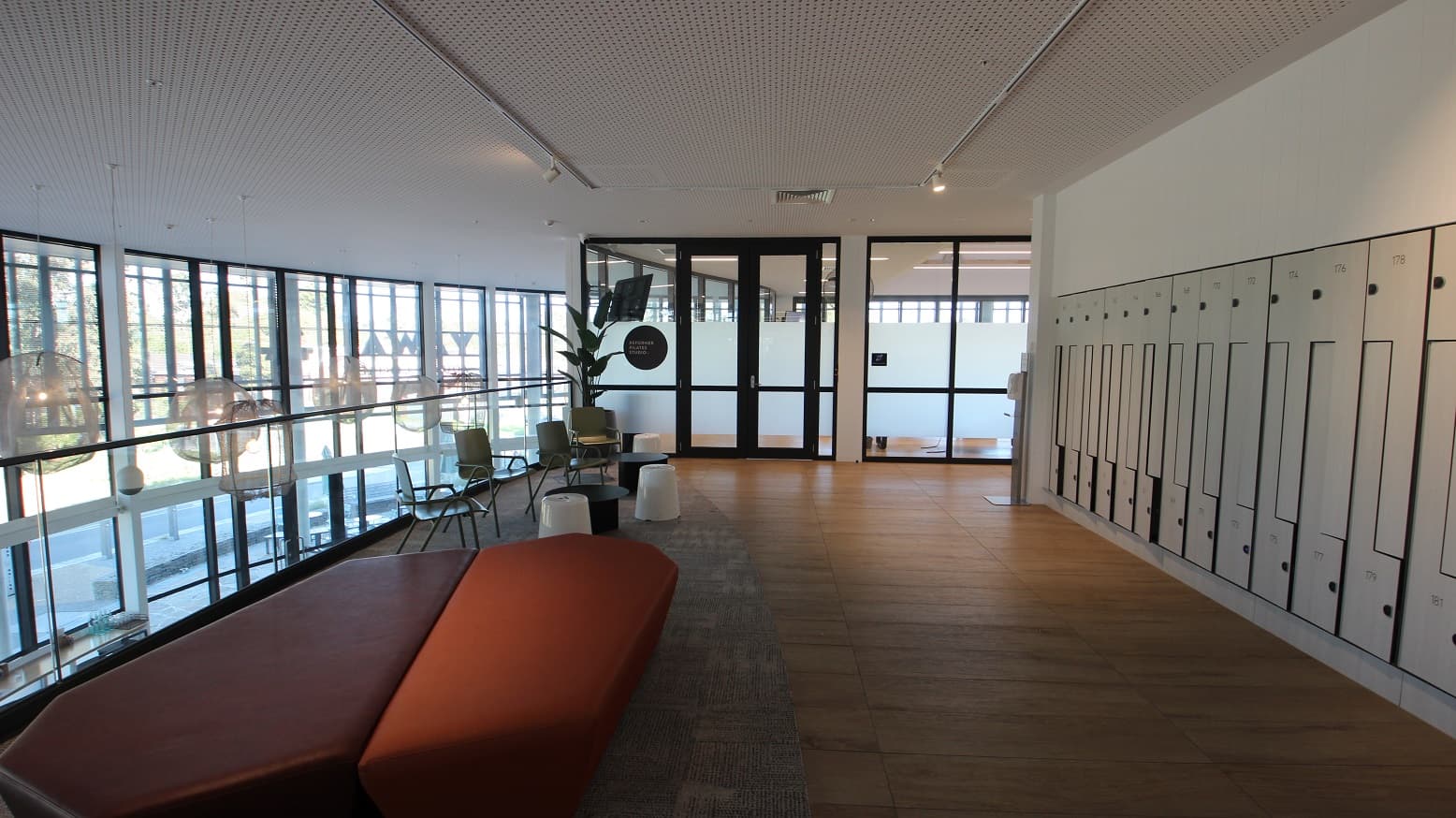 Locker area at Yawa Aquatic Centre. Each locker has designated numbers on top and below. This has an overlooking view of the Elements Eatery on the ground floor as seen by the jelly-like lighting hanging on the ceiling. There are also orange and red sofa chairs that have irregular shapes and green armchairs with black coffee tables. The seating area has a grey finish.