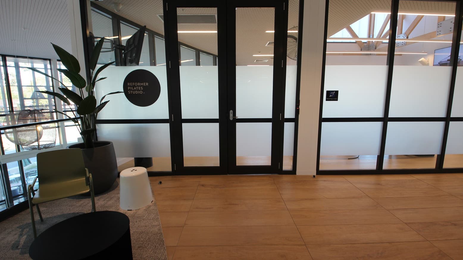 A closer look at the entry doors to the Rerformer Pilates Studio. There is a large potted plant on the left side, next to a green armchair, and within the grey floor mat. The studio can be identified by the black circular signboard on the left glass door.