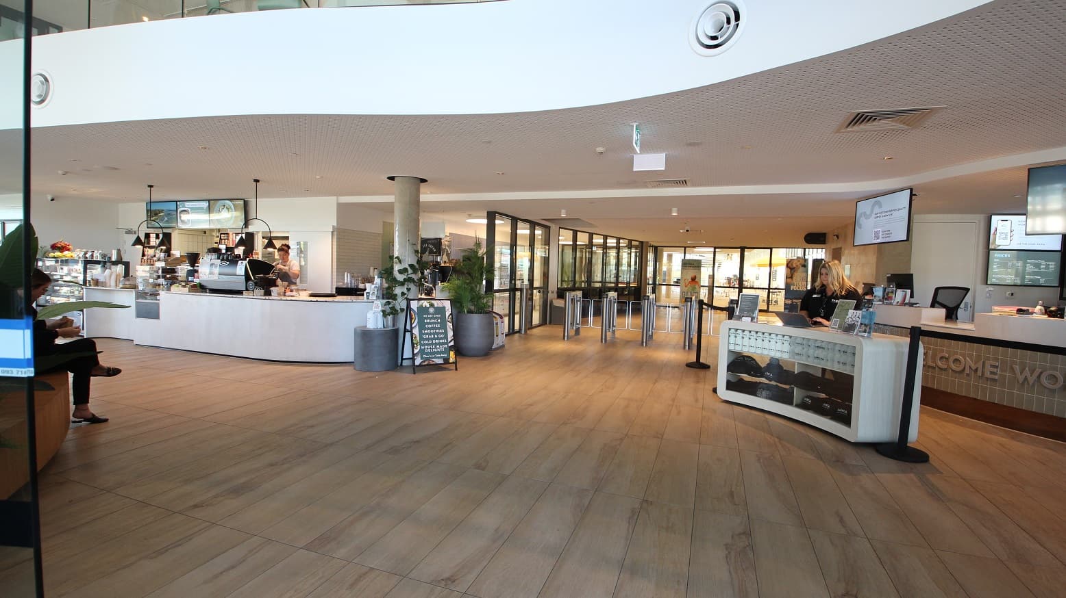 Upon entering the motion-censored glass entrance doors, there is a Café to the left, and the reception and retail areas are to the right. Near the Café counter is a glass display showcasing an array of pastries. The floor is made of ash hardwood, while the curvy ceiling is white with noticeable aesthetic holes as designed. The facility is well-lit due to the natural lighting filtering through the glass walls.