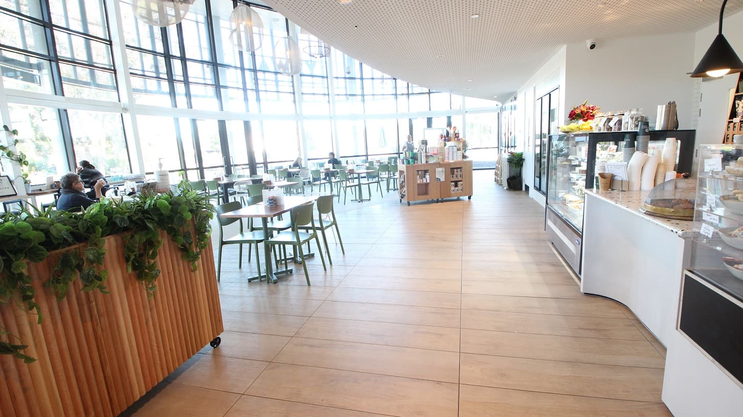 The café's seating area is illuminated by natural light coming in through the glass walls, and jelly-like artificial lights hanging from the ceiling provide additional lighting support. The seating area is composed of wooden tables, green and white chairs. There is a wooden planter box with artificial plants that has wheels that make it easy to move around.