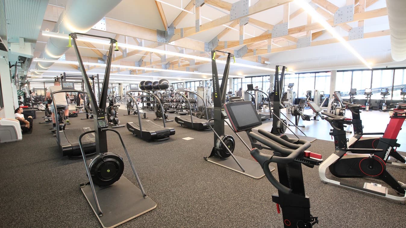 There is also a Skierge rowing machine, gym balls, curve treadmills, kettlebell swing, and exercise bikes inside the gym at Yawa Aquatic Centre. This section has a grey rubber mat floor. The industrial roofing frames that are a combination of wood and metal are usually found in barns or farms and the long string of artificial light connected to the opposite wall can be seen in this image.