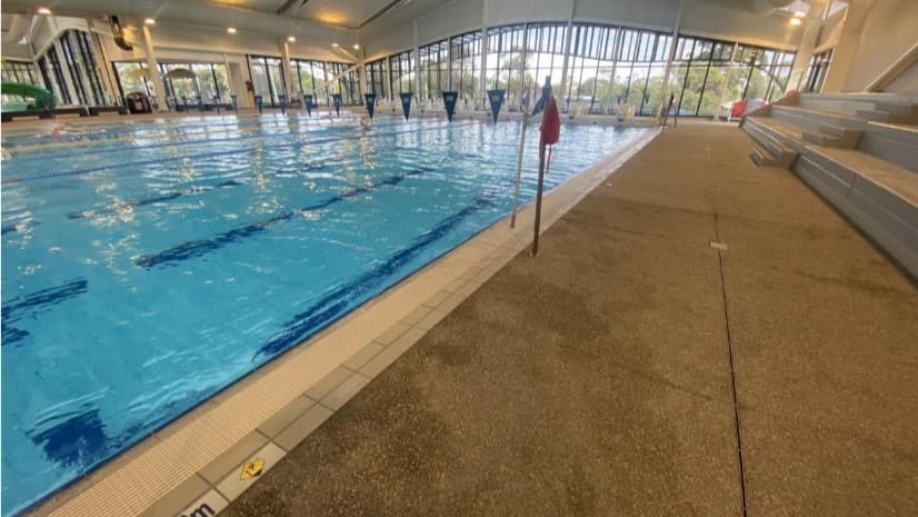 On the left, a pool with multiple lanes, delineated by floating ropes, is observed. Triangular banners hang across the pool. On the right, a pool deck with tiered seating is visible. Glass walls enclose the area.