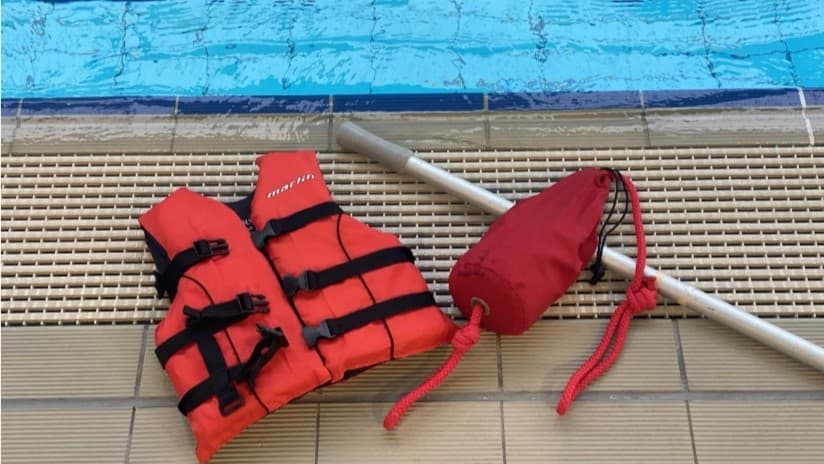 A red life jacket and red bean bag rest on the pool overflow grid beside a swimming pool. The bean bag has a rope attached. A metal pole with an anti-slip handle is positioned alongside the bean bag.