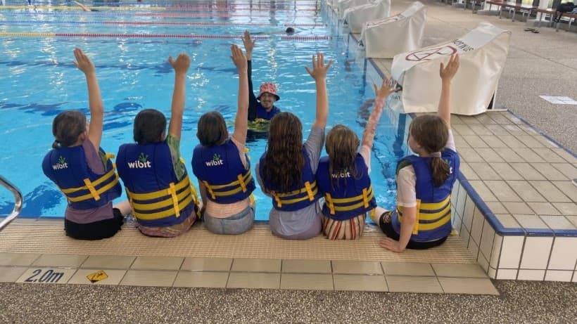 This image shows a group of children sitting at the edge of a swimming pool. They are wearing purple t-shirts and yellow life vests. The kids are seated with their backs to the camera, facing an instructor who is in the pool wearing a red cap and goggles. The instructor is also raising their hands, and the children are mimicking the gesture. A raised pool deck with diving boards is positioned on the left.