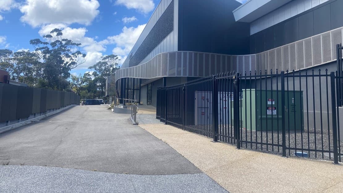 A partial view of a building with a grey concrete facade with a glass door with a black arched metal canopy. The foreground features a concrete ramp with metal railings, leading to the doors. The floor is covered in light and dark grey-speckled tiles. Wild grass is visible on the left.