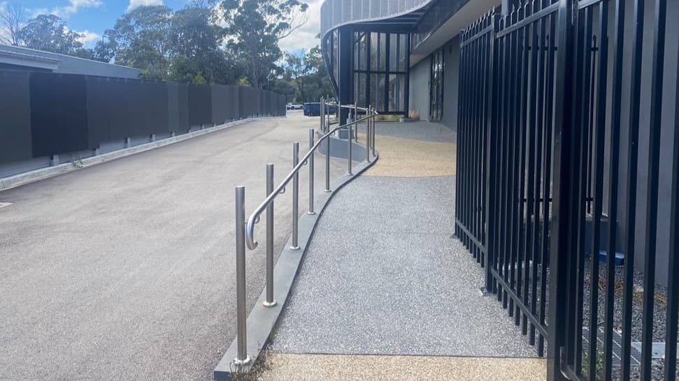 To the left, a black metal fence aligns with a concrete pathway bordered by metal handrails. On the right, a tarmac car park stretches into the distance. Adjacent to the car park is a concrete ramp with handrails leading towards the building. The modern structure boasts curved metal panels and large windows. Trees provide a backdrop in the background.