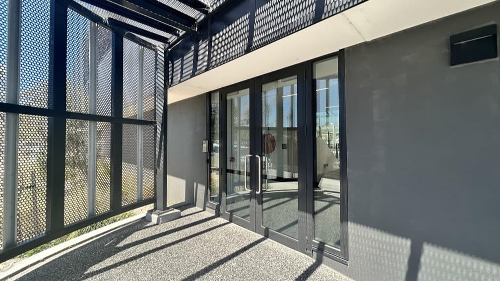 A modern building entrance featuring double glass doors with metal handles, flanked by black, perforated metal panels. The structure is complemented by a black metal framework and is well-lit by natural light. The paving is a textured grey surface.