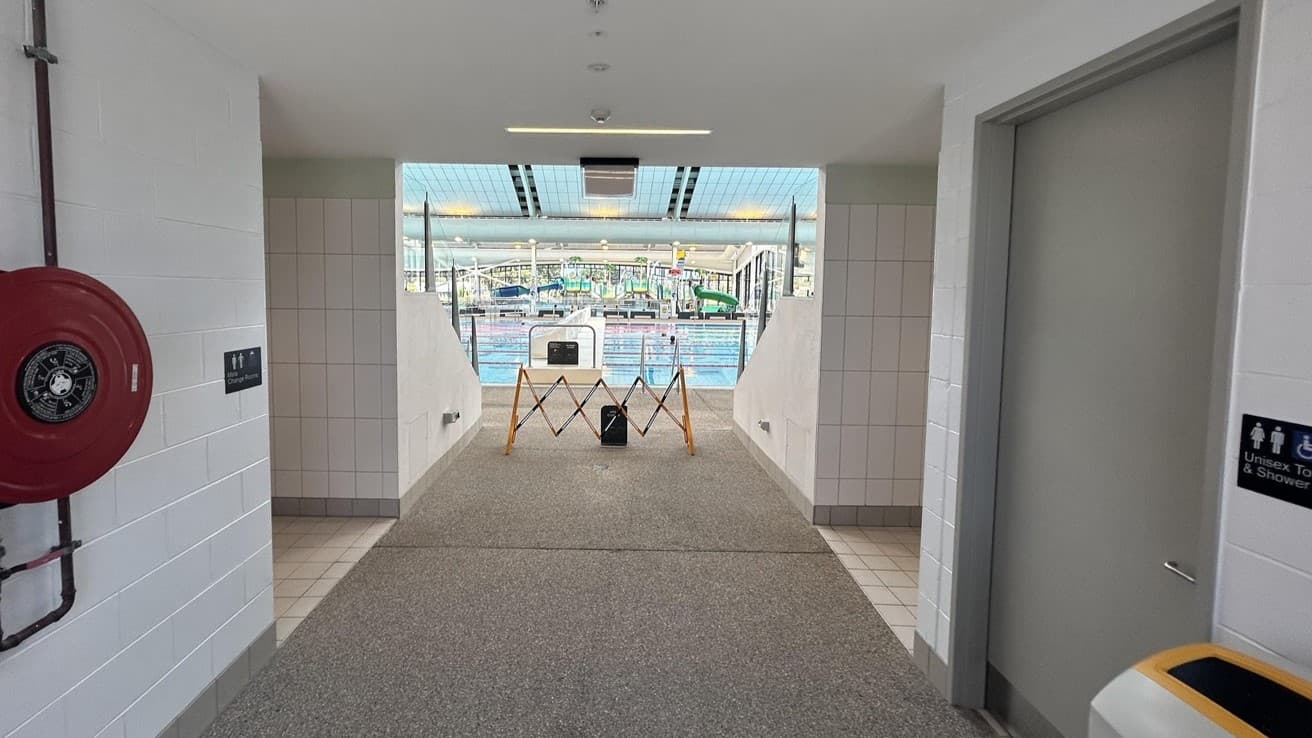 A corridor with white tiled walls and a speckled grey floor leads to a swimming pool. On the left, a bathroom sign is affixed to the wall. A firehose reel hangs nearby. On the right, a door leads to a bathroom, marked with a sign. In front is a bin. A retractable barrier is positioned in the middle of the pathway. Fluorescent lights illuminate the ceiling, and a retractable barrier partially blocks the doorway.