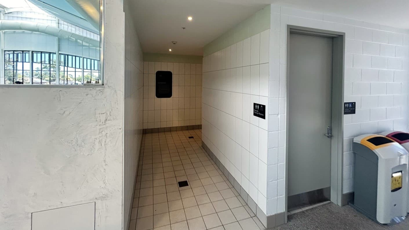 To the left, a corridor leads to the toilet, with signage affixed to the white tiled wall. The floor is tiled in light grey. Overhead, white ceiling panels continue throughout. To the right, a door leads to the toilet. Rubbish bins are placed against the wall.