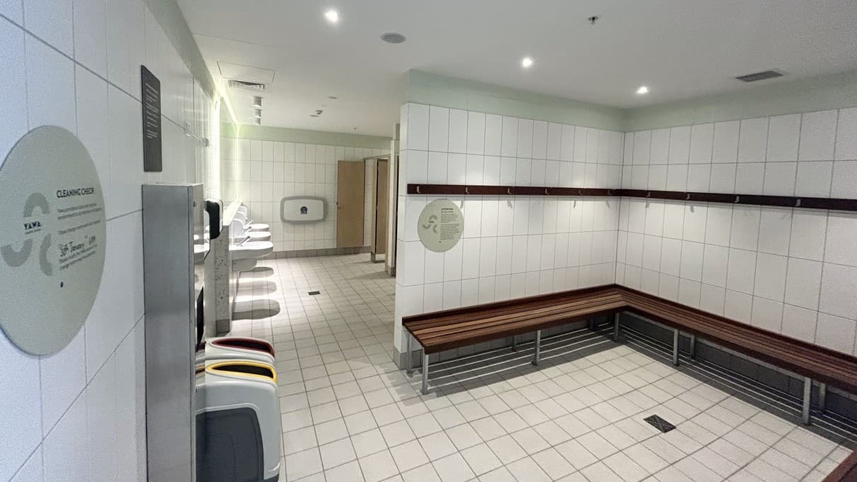 The interior shows a bathroom or changing room with white tiles on the walls and floor, creating a bright and sterile environment. On the left, a large circular sign displays facility rules. Wash basins and a foldable change table are fixed to the walls. To the right, wooden benches form an L-shape along the tiled walls. Further in, bathroom stalls are visible. Rubbish bins are positioned against the wall.