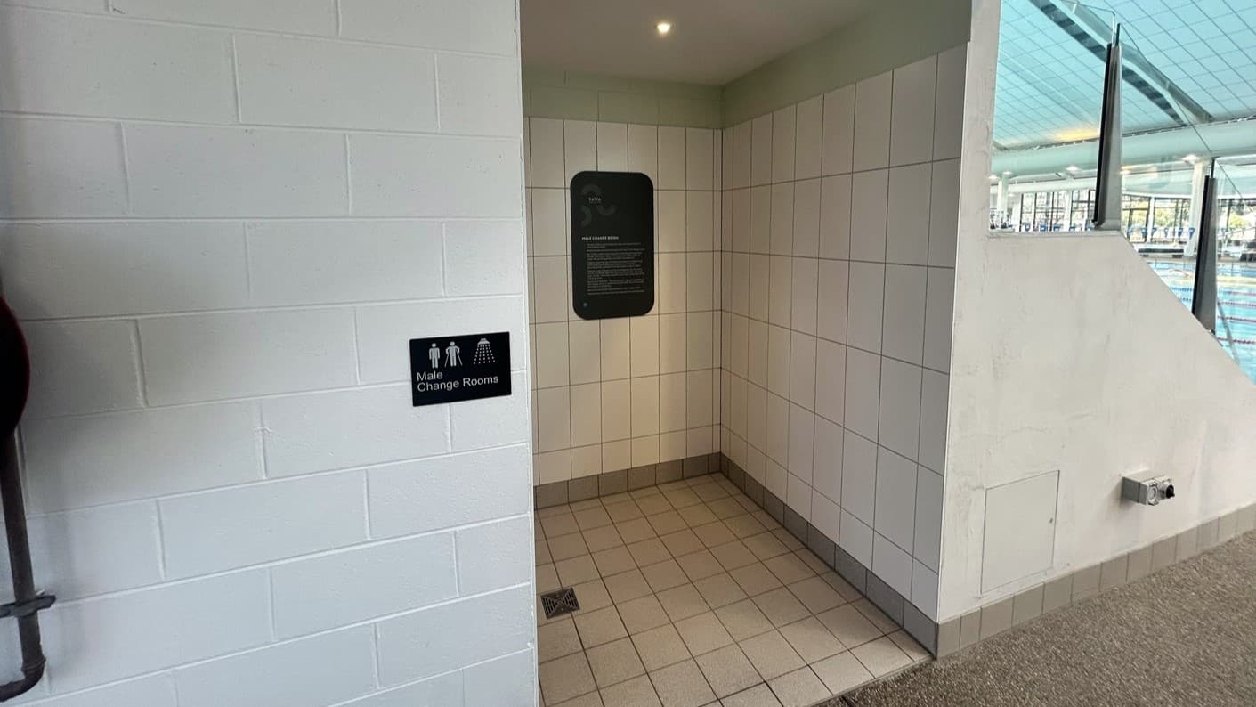 The Male Change room displays white and blue wall tiles with signage affixed. A partial view of the pool is on the right. The floor is concrete, and the ceiling is white with exposed aircon ducts and metal beams.