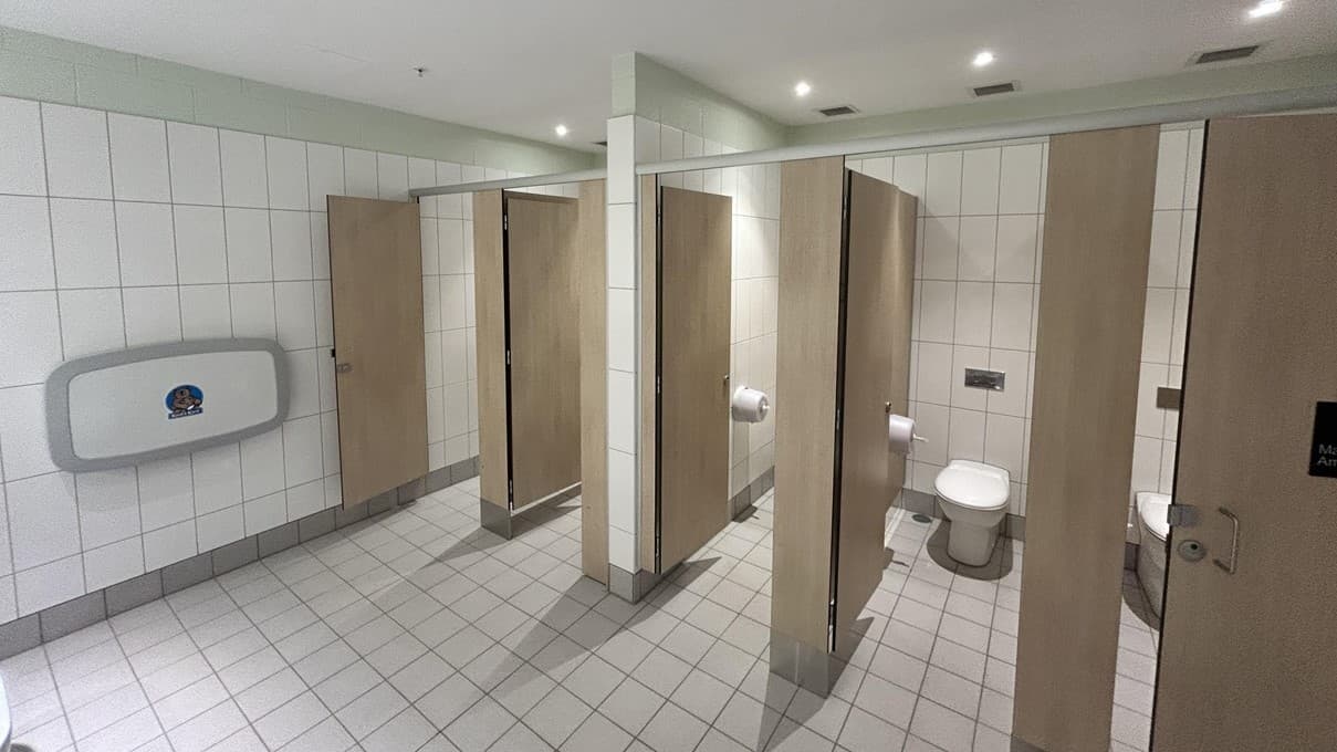 Inside the accessible bathroom, multiple bathroom stalls with brown doors are visible, one slightly ajar revealing a toilet inside. To the left of the stalls, an accessible toilet stall appears larger with supporting handrails and a visible toilet. Far left, a wall-mounted baby changing table is observed. White-tiled floor and walls complement the white ceiling with recessed lights. Signage on one stall door indicates accessibility features.