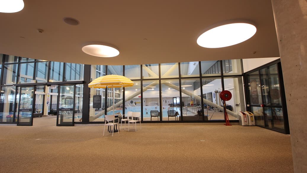 A yellow umbrella with blue tables and white chairs creates a light summery feel inside the aquatic facility of Yawa Aquatic Centre. This section of the facility has a granite floor with a curvy ceiling with large circular fluorescent lights. On the right side of the glass wall is a roll of fire hose and two rubbish bins with a unique design.