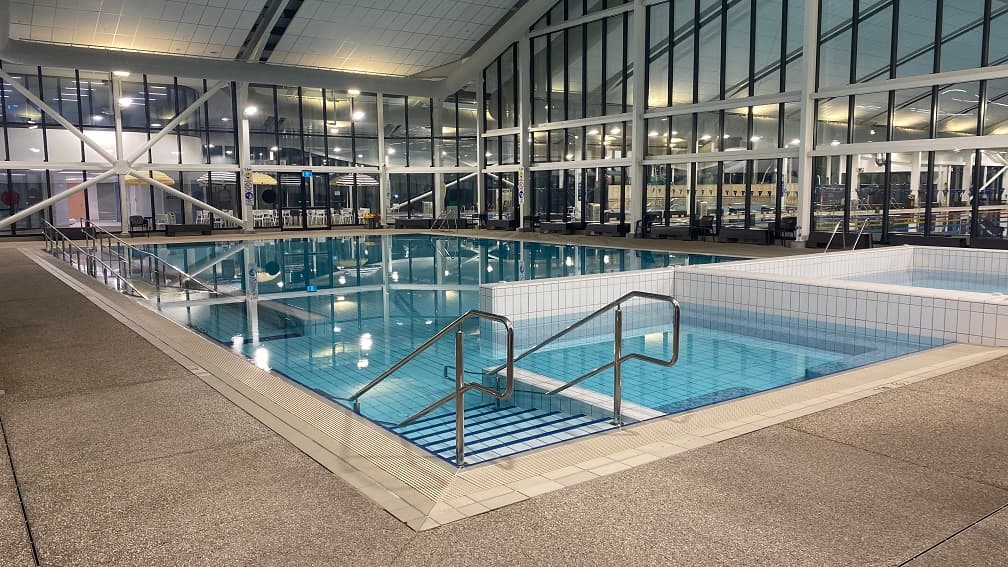 There are stairs leading into the warm water pool directly opposite the ramp entrance. The glass walls have unique cross frames. This position has a view of the series of chairs and tables close to the change room and the learn-to-swim pool through the glass walls. Lights coming from artificial light bulbs are reflected on the pool water. There is a brown tile that separates the pool from the granite floor.
