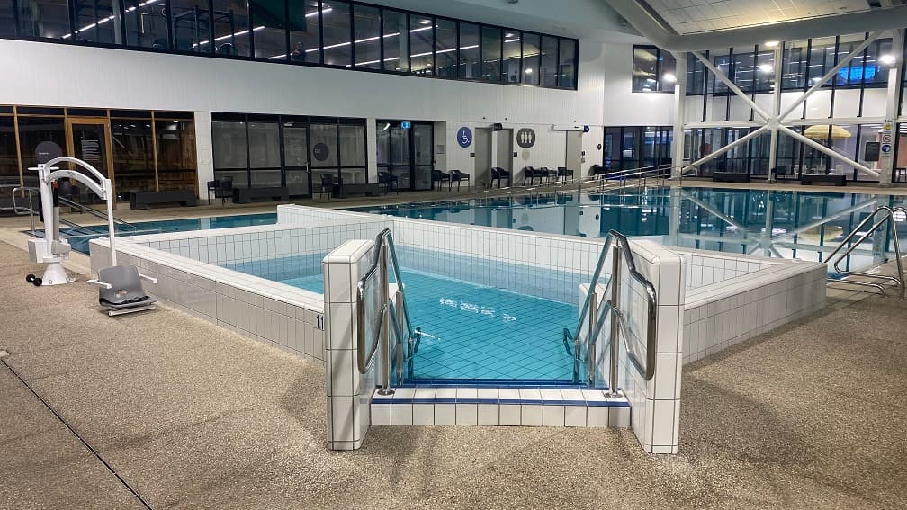 Stairs leading to the rectangular spa connected to the warm water pool. The steps have blue stripes on their edges. On the left side is a white pool hoist with a grey seat. Black chairs and benches can be seen on the sides and near the accessible bathrooms. The second floor is identifiable through the glass windows with black metal frames. Different pool warning signs can be found on one of the structural posts in this facility.