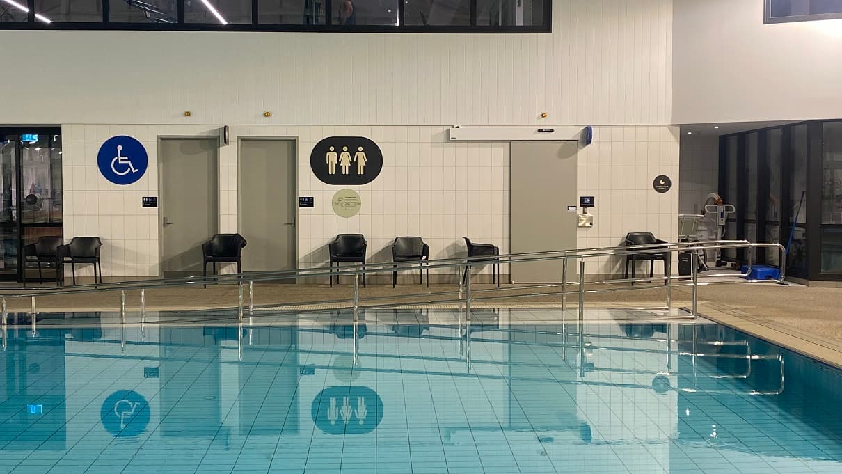 Accessible bathrooms can easily be distinguished by the large sign boards placed on the upper right wall of the grey doors. A Blue accessible signboard can be seen on the left while a black circular signboard with three icons for different genders (male, female, non-binary) can be seen on the right. Near the bathrooms are black chairs. On the open space on the right is another pool hoist reserved for future occasions.