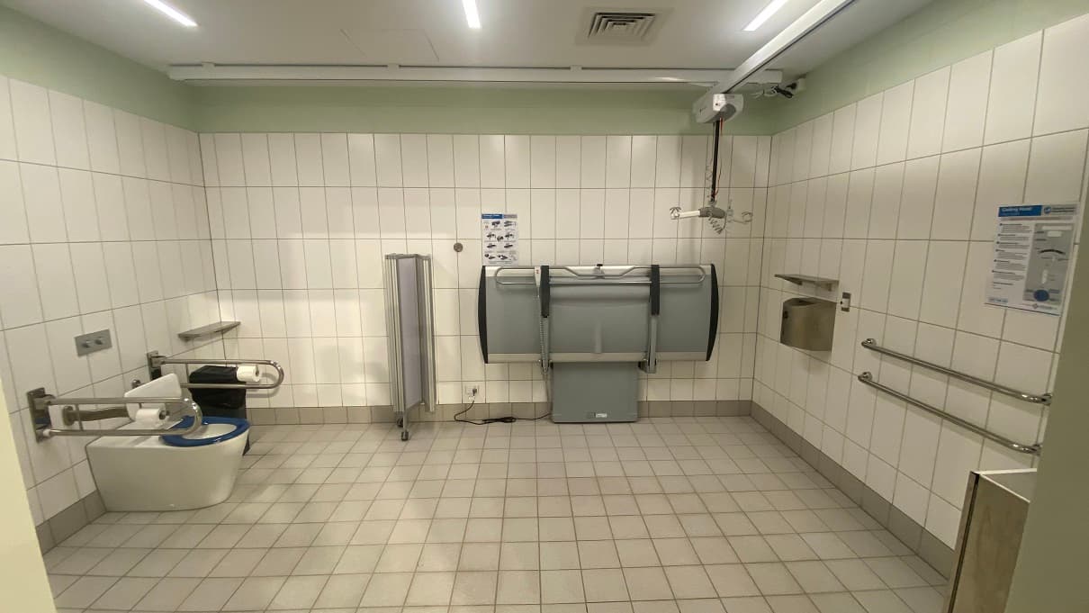 Inside the accessible bathroom of the Warm Water pool at Yawa Aquatic Centre. The bathroom has a white tiled wall with a lime green portion before the ceiling. There is a white and grey portable hoist inside, a grey adult changing table, a movable panel for privacy, grab bars mounted on the wall and on the sides of the toilet area. The accessible toilet has a blue toilet cover and white back cushion for support.