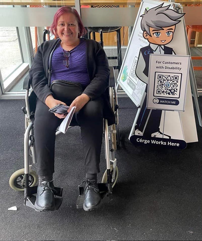 A woman in a wheelchair beside a Cérge cartoon cutout with a QR sign. A woman in a wheelchair beside a Cérge cartoon cutout with a QR sign.
