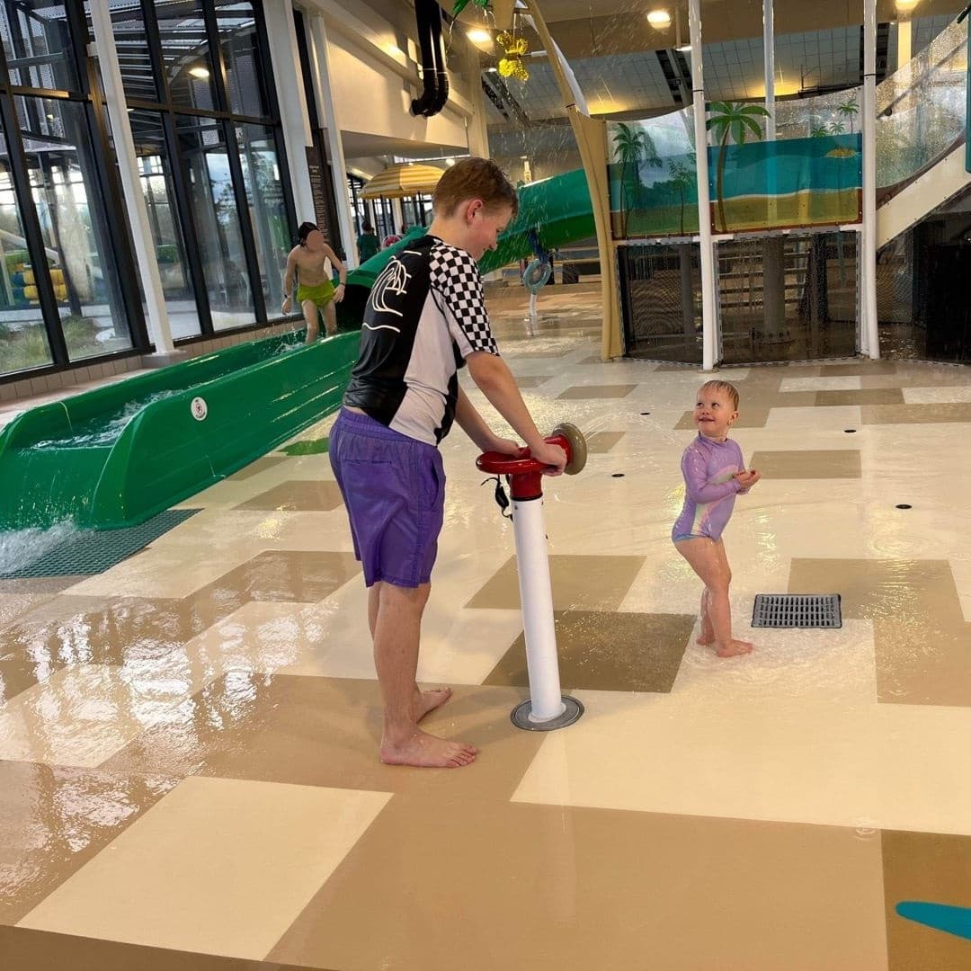 A boy helps a toddler at a water play area with a splash pad. A boy helps a toddler at a water play area with a splash pad.