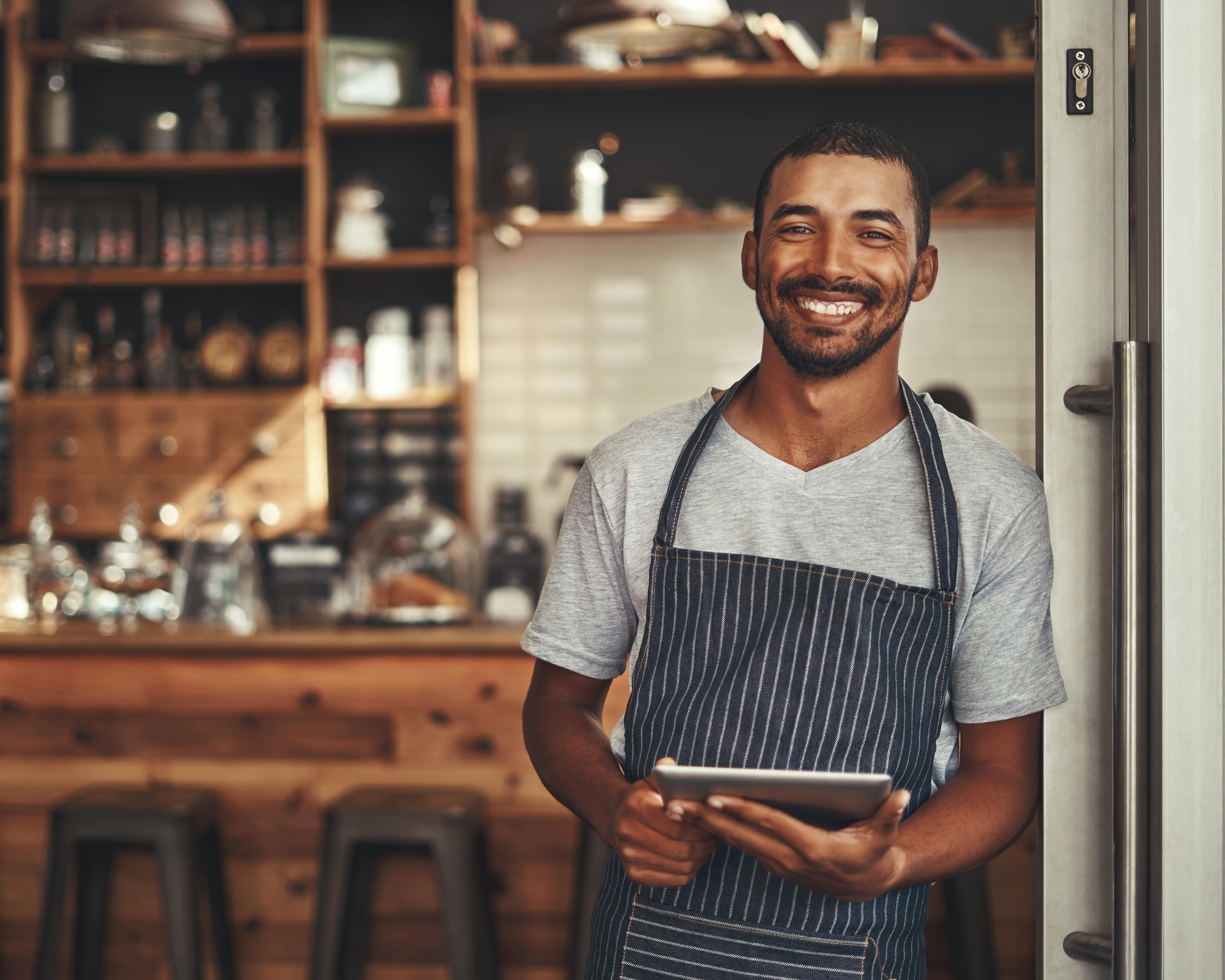 A young waiter of a cafe or restaurant meeting you as you arrive. He has a big bright smiling face and holding a digital tablet. A feeling of happiness and fun greets you for great customer service. The background is a warm cafe setting with lots of dark coloured timbers.
  Email alttext@cerge to suggest improvements to our alt text