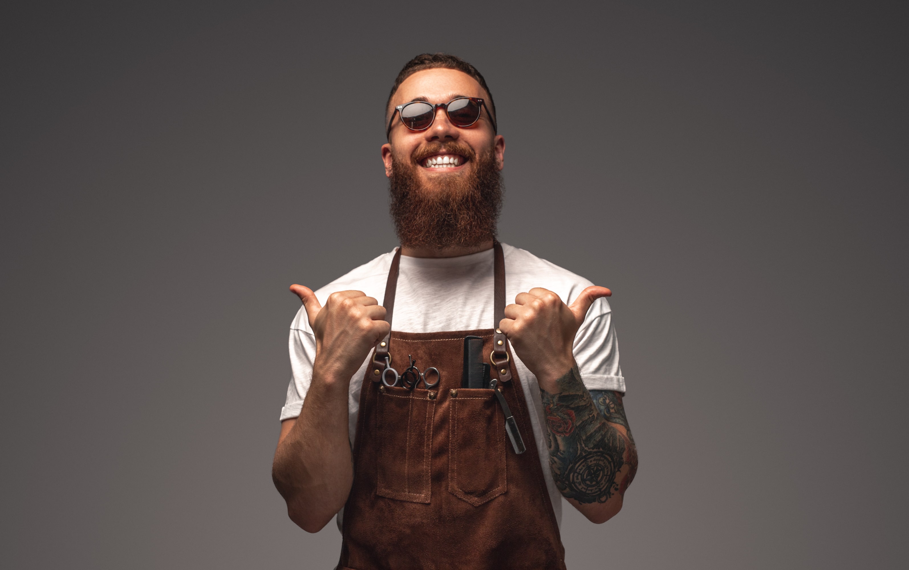 A totally hip barber with one of the biggest smiles ever.  With quite a long but well-trimmed beard, neatly clipped hair and small oval sunglasses he has a fun vibe.  He is wearing a leather apron with a few pairs of scissors in the chest pockets and giving two big thumbs up.  The background is a plain grey colour without any features. To enhance our alttext, email alttext@cerge.app with suggestions.