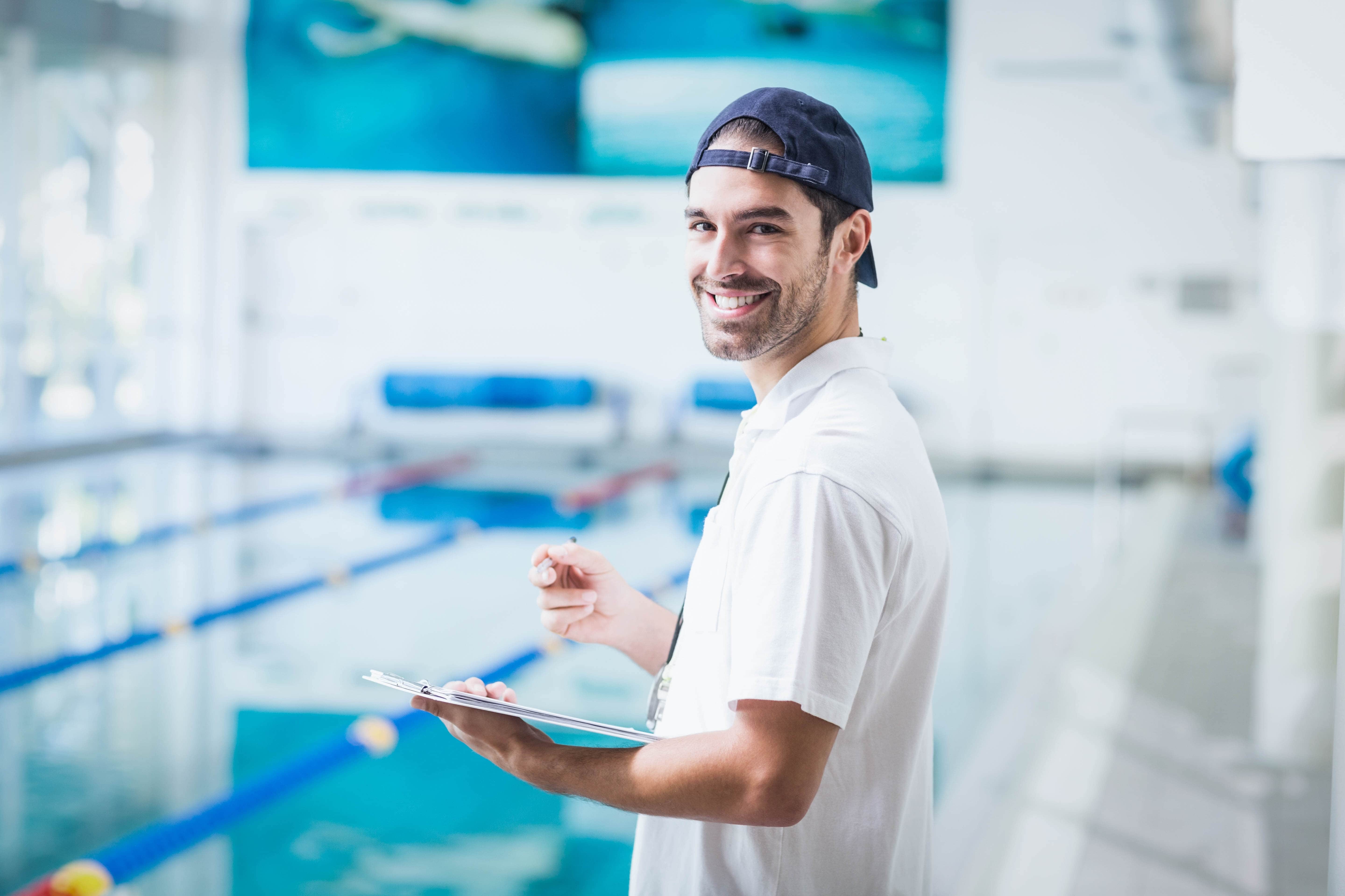 A male swimming instructor is standing and smiling toward the camera whilst holding a clipboard and a pen to take notes.  A 25-meter indoor pool is in the background.  The swimming instructor has his hat on backwards and with his casual facial expressions he looks very approachable and friendly. To enhance our alttext, email alttext@cerge.app with suggestions.