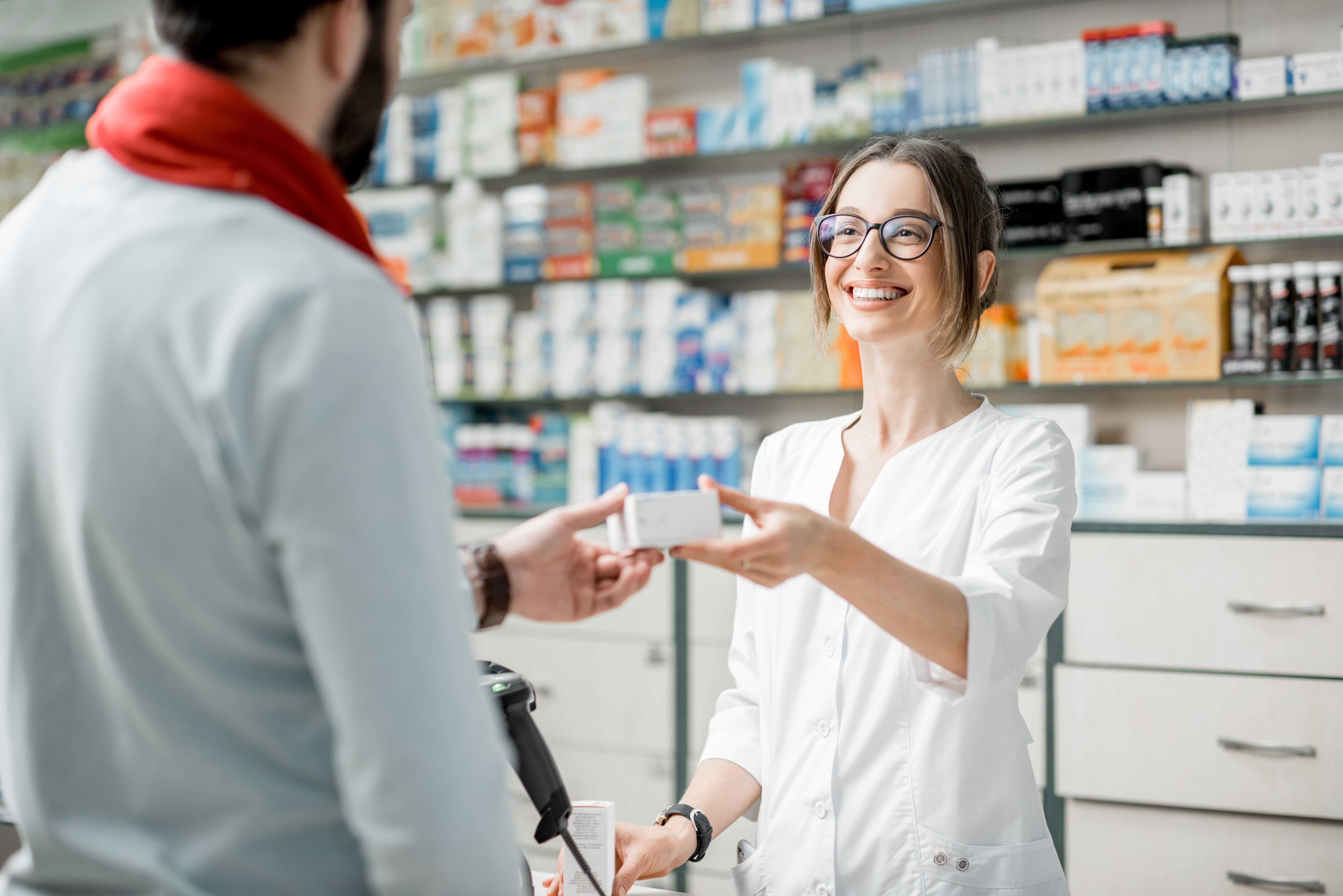 A female pharmacist is handing over some medicines to a customer within her store.  Her bright smile and positive body language infers the experience was fun and uplifting for both her and the customer.  She has a typical white pharmacist top on with the background full of other medicines.
  To enhance our alttext, email alttext@cerge.app with suggestions.